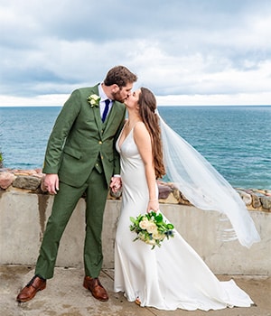 A bride in a sleek white gown and a groom in a green suit kissing by the ocean.