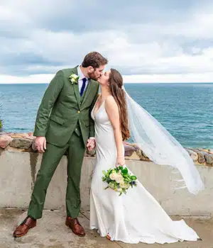 A bride in a sleek white gown and a groom in a green suit kissing by the ocean.